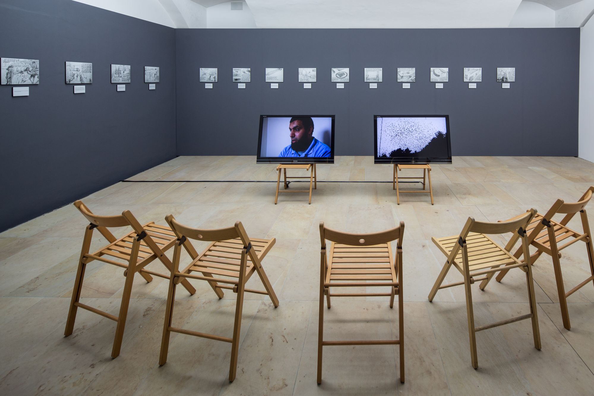 Ein Foto aus der Ausstellung. An den Wänden hängen Zeichnungen. Im Raum stehen zwei Stühle mit zwei Fernsehern. Ein Halbkreis aus Stühlen steht um die Fernseher herum. 