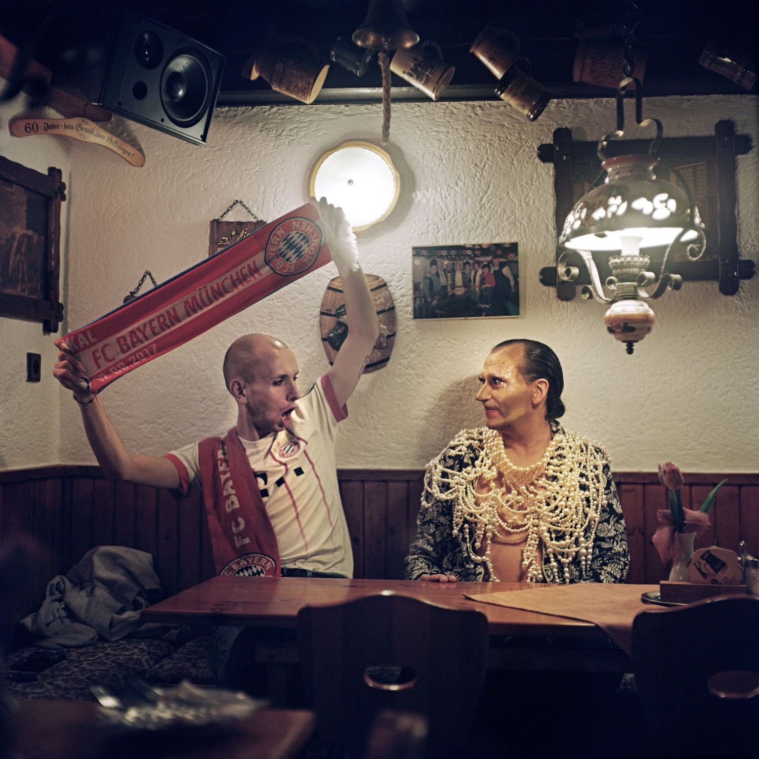 A man wearing a soccer jersey and a man wearing pearl necklaces are sitting in a bar