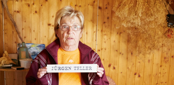 Ältere Dame vor einer hölzernen Wand, die ein Schild mit dem Namen Jürgen Teller hält