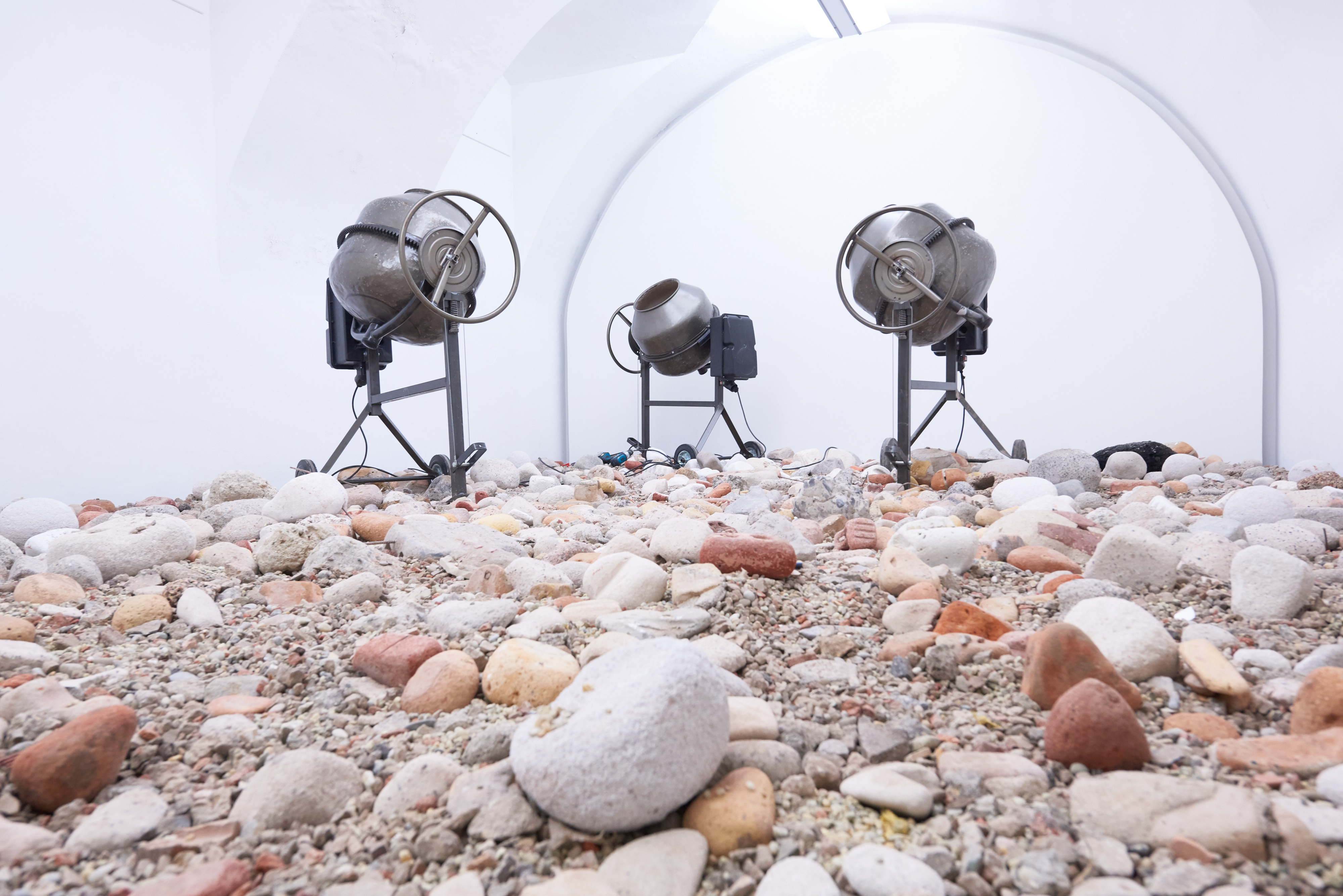 View of the work Clockwork by artists Julius von Bismarck, which consists of three concrete mixers that incessantly hurl stones. These stones cover the entire floor.